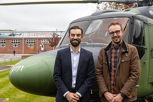 Adam Dance MP and Ben Clarke from Unite stand together in front of a military helicopter at the Leonardo facility. Adam wears a brown jacket and plaid shirt, while Ben is dressed in a navy suit with a white shirt. The Leonardo logo is visible on the building in the background.