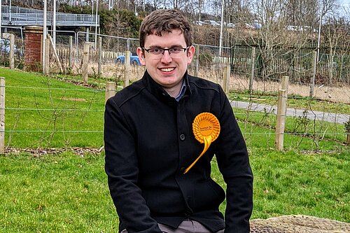 Grant Toghill sitting on a wall wearing a Scottish Lib Dem rosette.