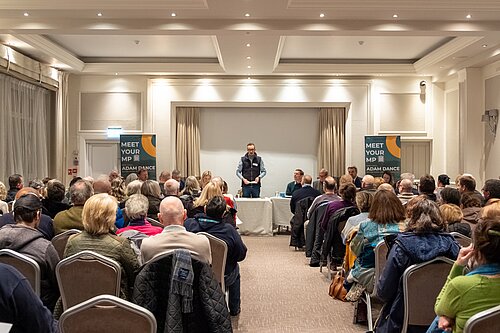 Adam Dance MP stands at the front of a well-attended public meeting, addressing a seated audience in a conference room. Behind him, a panel of individuals is seated at a table, and two banners read "MEET YOUR MP - ADAM DANCE." The room is filled with people listening attentively.