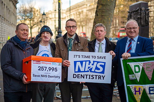 Group photo featuring Tim Farron, Adam Dance, Ian Roome and James MacCleary and a campaigner, holding signs and a petition box displaying '257,995 people say Save NHS Dentistry,' with London streets and iconic architecture in the background.