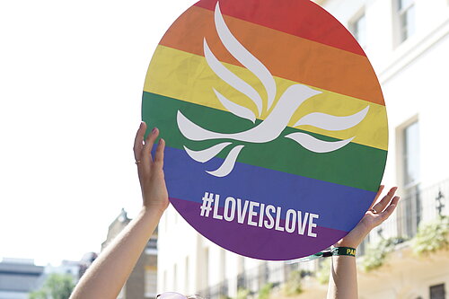 Young person holding a round sign over their head with a rainbow-striped, pride flag design overlaid with the Liberal Democrat bird of liberty emblem and #LoveIsLove