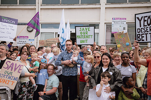 Adam Dance MP stands holding a baby among a large group of parents, children, and supporters outside a hospital. They are holding handmade signs that say things like "Save Our Maternity Unit" and "Yeovil Deserves Better," as part of a protest to protect local maternity services.