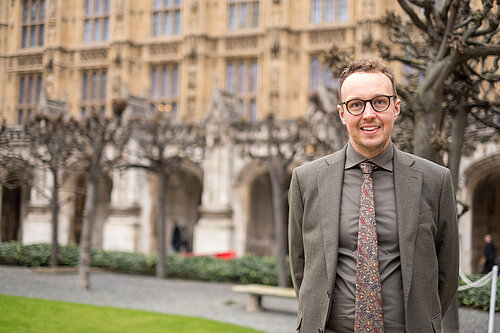 Adam Dance MP standing outdoors in front of the historic architecture of the Houses of Parliament. He is wearing a grey suit, a patterned tie, and glasses, smiling at the camera. The background features leafless trees and a well-maintained garden area.